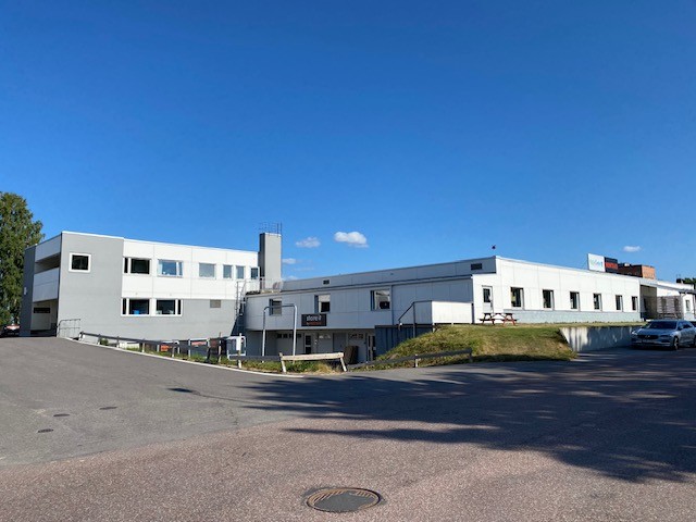 Vit och grå byggnad i olika nivåer. I förgrunden väg och parkering.