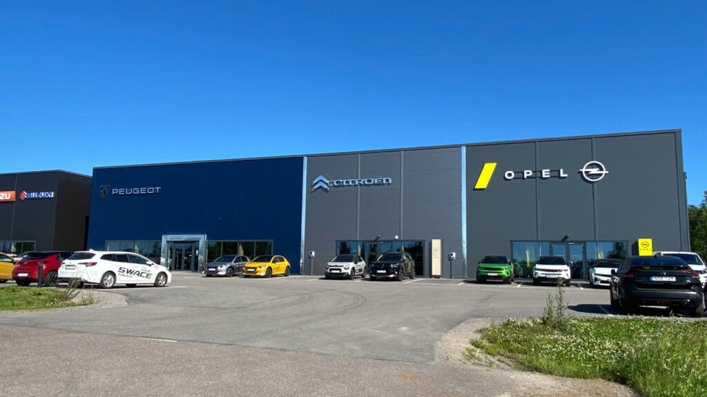Grå fyrkantig byggnad med bilmärken/loggor på fasad. Framför byggnaden står det flera olika bilar uppställda